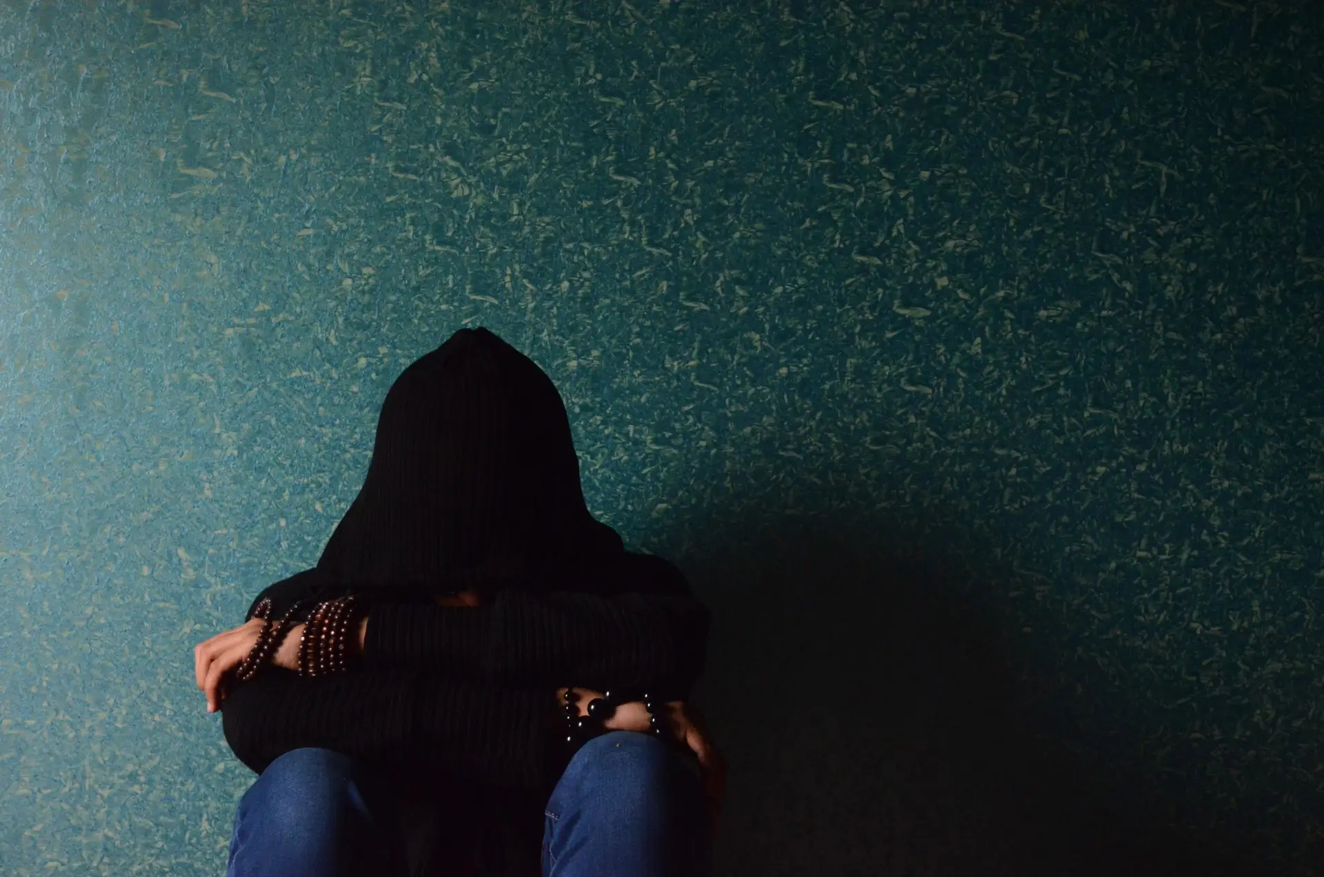 person covering face with hood sitting against a wall