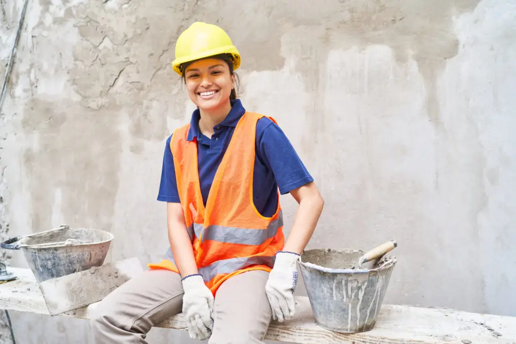 A woman smiling with a yellow hard hat and an orange vest sits on a board in front of a concrete wall she has just finished patching. This represents how to increase dopamine by completing a task.