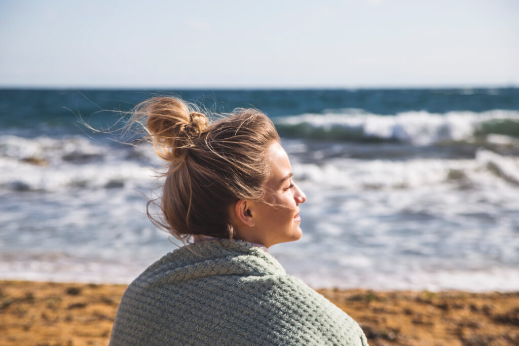A woman sits at the beach in a blanket watching the waves roll in, which is a relaxing vagus nerve exercise.