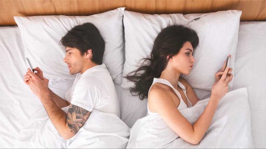 A couple in bed, on their phones, engaging in conflict avoidance.