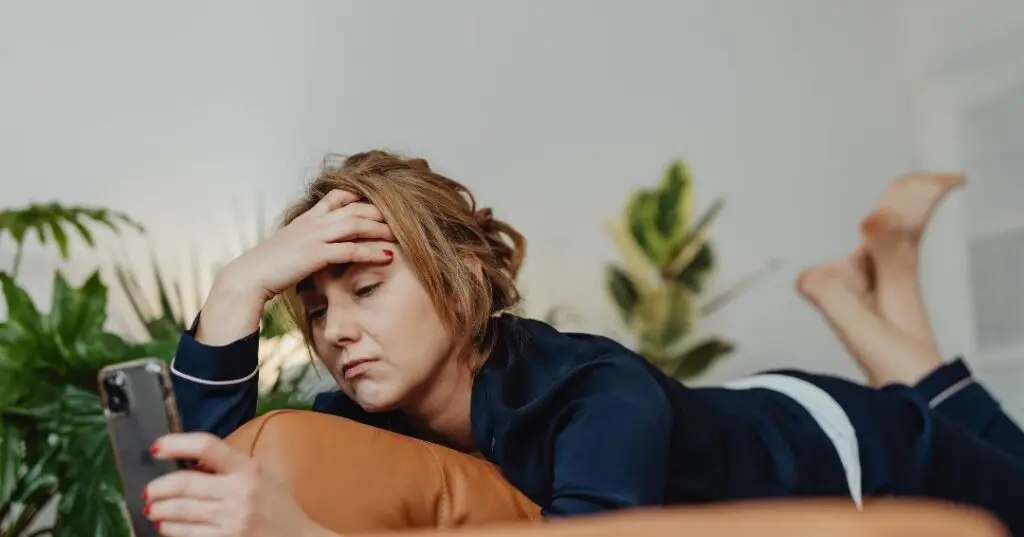 A woman laying on her stomach scrolls mindlessly through her phone illustrating how one of the symptoms of depression is that it can cause you to feel unmotivated.