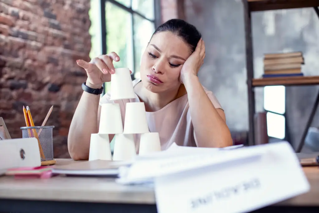 7 Signs of High Functioning Anxiety: Plus How To Relieve Some Of That Anxiety A woman is stacking cups in a pyramid on her work desk showing how procrastination is a part of high functioning anxiety.