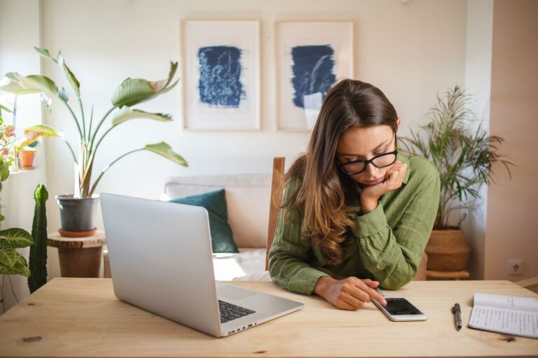 Young woman distracted by her phone when she should be working on her laptop, because she is struggling with pathological demand avoidance.