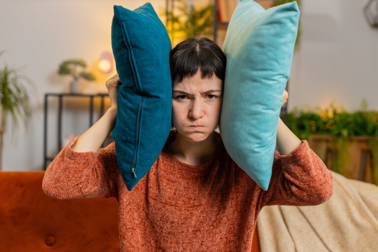 Woman sitting in a living room, looking frustrated as her head is squished between two pillows, illustrating the concept of overstimulation meaning and sensory overwhelm.