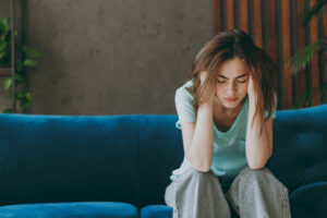 A young woman sits on a blue couch with her head in her hands, eyes closed, looking distressed, representing the emotional weight and self-critical thoughts common in self deprecating behavior