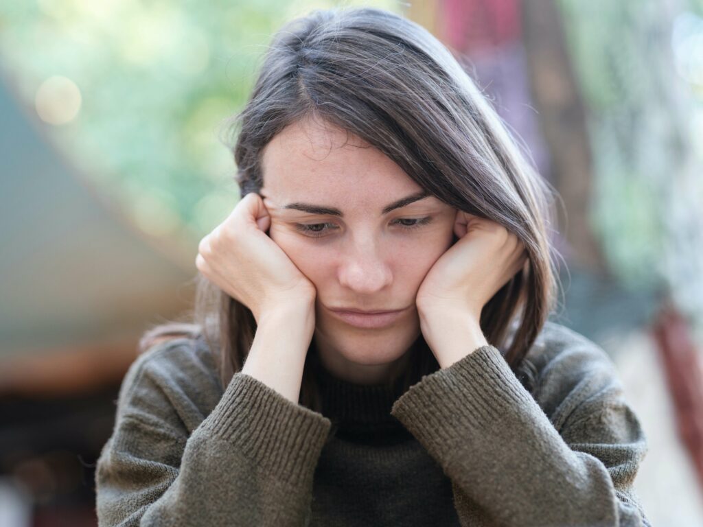Woman sitting with her head resting in her hands, looking down with a sad expression, reflecting the emotional impact of self deprecating beliefs and low self-worth.
