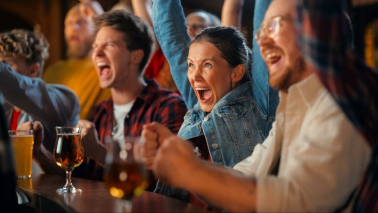 A group of friends cheering loudly at a bar with raised arms and intense expressions, illustrating deindividuation as individuals become absorbed in shared emotion and group behavior.