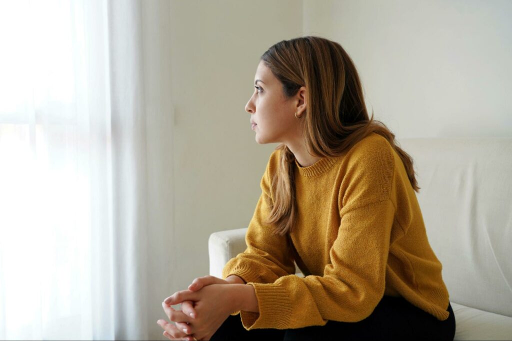 A woman sitting alone indoors, gazing thoughtfully and sadly out a window, feeling conflicted.
