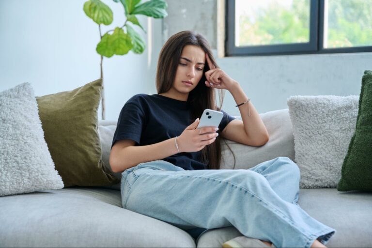 Young woman sitting on a couch looking thoughtfully at her phone, appearing concerned or overwhelmed.
