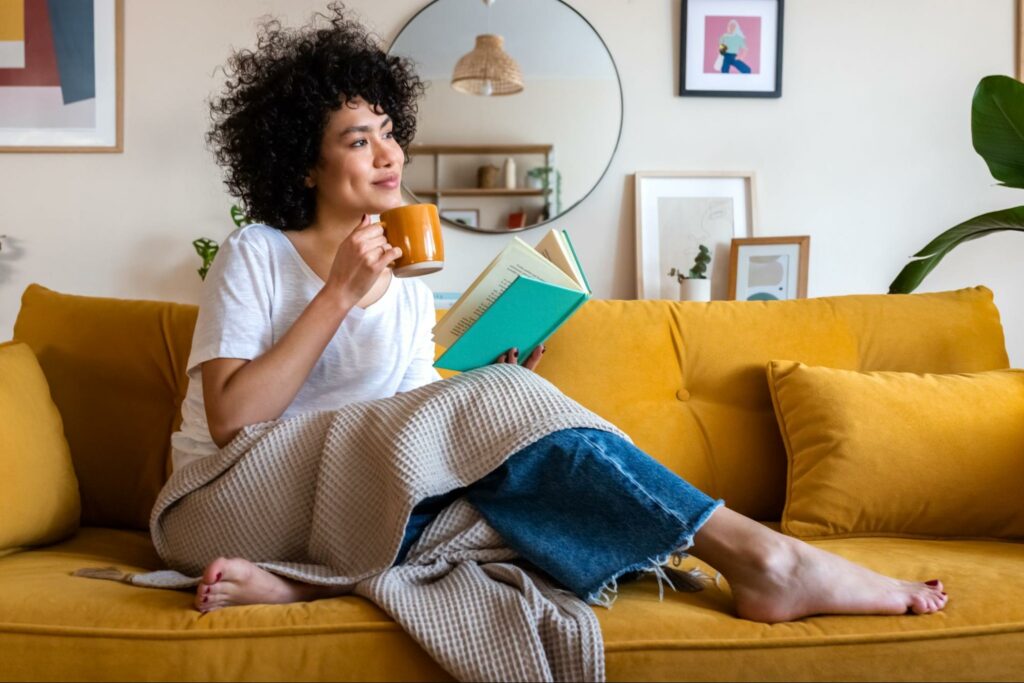 Young woman sitting comfortably on a couch, reading a book and holding a mug while appearing calm and relaxed in a cozy home setting.
