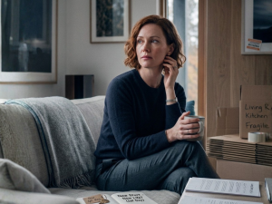 A pensive 35-year-old woman sitting on a gray sofa in a modern home, holding a mug and looking thoughtfully out a window. Around her are signs of a major life transition, including packed moving boxes labeled "Living Room" and "Kitchen," documents on a coffee table, and an open journal with a set of keys.