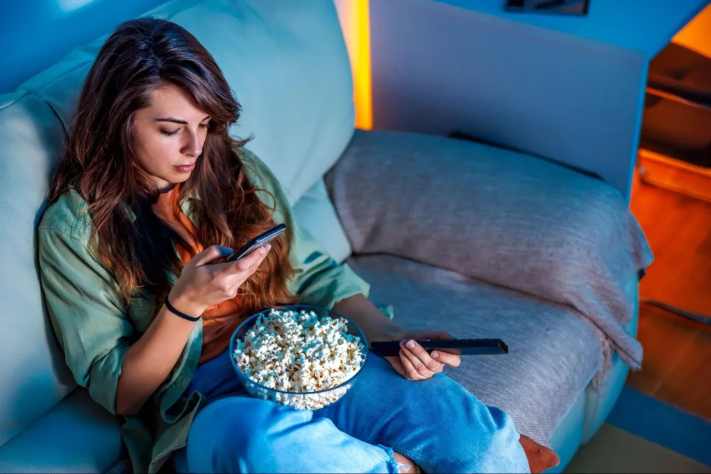 A woman is shown scrolling on her phone while simultaneously looking for something to watch on television.