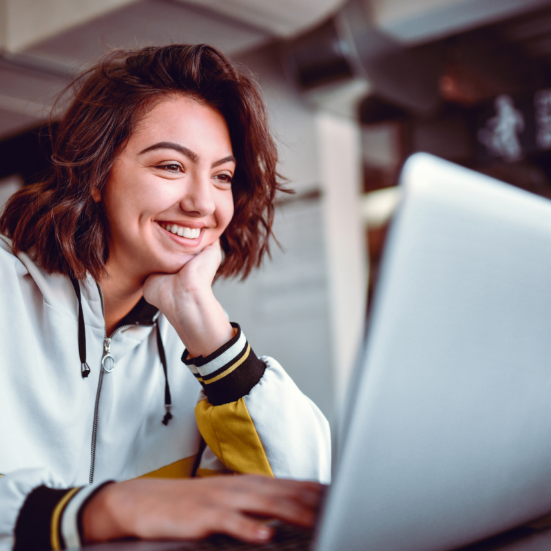 A young woman smiling as she looks at her laptop screen in a modern indoor setting, suggesting a positive and engaging virtual experience- 3 tips for starting online IFS therapy