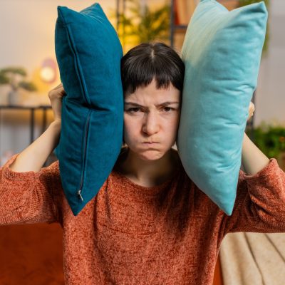 Woman sitting in a living room, looking frustrated as her head is squished between two pillows, illustrating the concept of overstimulation meaning and sensory overwhelm.