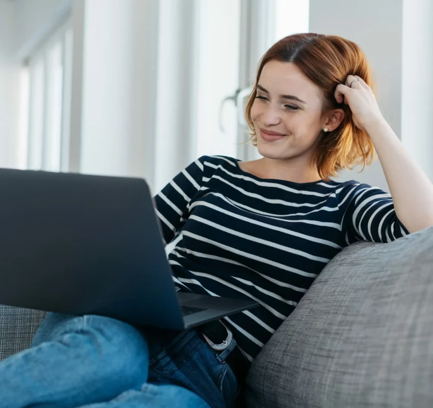 Cute young redhead woman with a lovely smile working on a laptop balanced on her knee as she relaxes on a sofa at home in a low angle view