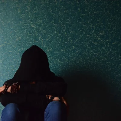 person covering face with hood sitting against a wall