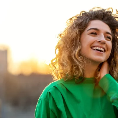 A woman in a green shirt stands in front of a building with her hand on her neck, smiling signifying that she has learned how to increase dopamine.