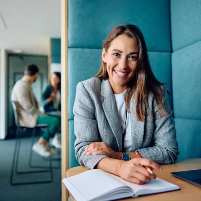 A photo of a smiling woman sitting at a table with a notebook. She is happy because she used her new executive functioning skills to be productive.