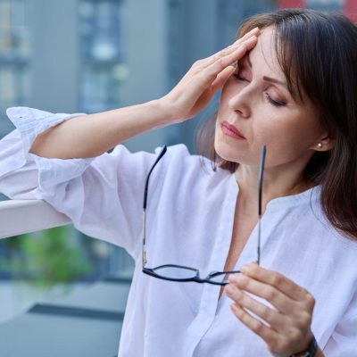 A woman holding glasses in her hand with her other hand on her head showing that she is anxious.