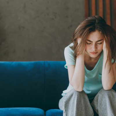 A young woman sits on a blue couch with her head in her hands, eyes closed, looking distressed, representing the emotional weight and self-critical thoughts common in self deprecating behavior