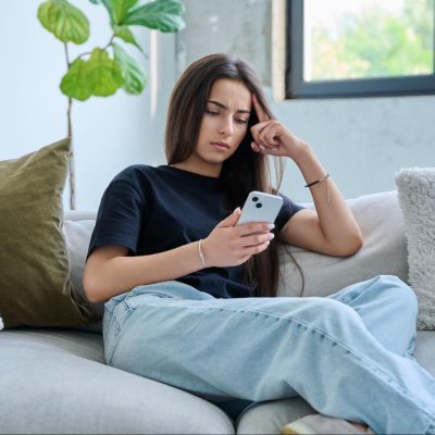 Young woman sitting on a couch looking thoughtfully at her phone, appearing concerned or overwhelmed.