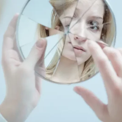 woman touching her reflection in broken mirror What no one tells you about body dysmorphic disorder . Makin Wellness . online body dysmorphia therapy
